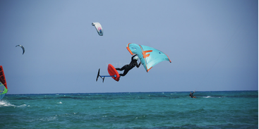 A person is wing foiling above turquoise ocean waves, holding a blue wing. Others are windsurfing and kite surfing nearby under a clear blue sky.