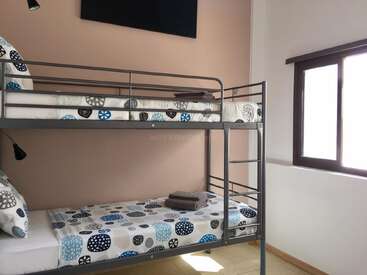 This image shows a tidy bedroom with a metal bunk bed, colorful patterned bedding, folded towels, a window with sunlight, and soft pink and white walls.
