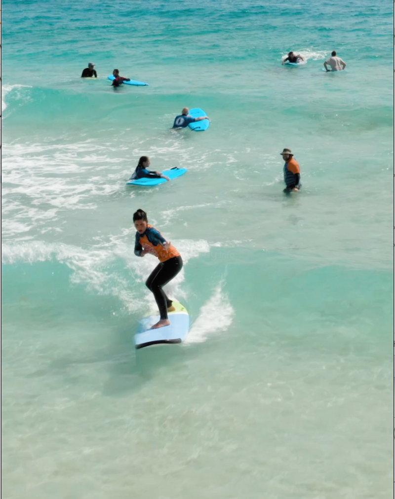 The image depicts a group of people surfing in the ocean, with one person prominently featured riding a wave on a blue surfboard.