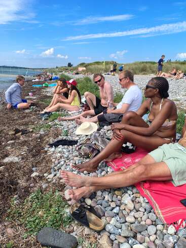 Un grupo de personas se relaja en una playa rocosa bajo un cielo azul brillante, charlando, tomando el sol y disfrutando de la compañía de los demás cerca del agua.