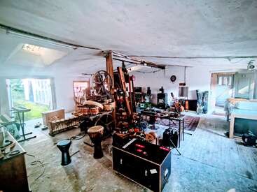 A spacious, rustic room filled with musical instruments, recording equipment, and eclectic furniture. Guitars, drums, keyboards, and various gear suggest it’s a creative music studio.