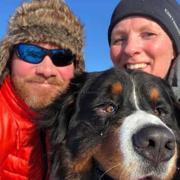 La imagen muestra a un hombre y una mujer posando con un gran perro frente a un cielo azul despejado en un día soleado, con ropa de abrigo de invierno.