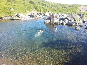 Children play in a clear, shallow river surrounded by rocks and greenery. Some swim and others stand on rocks, enjoying a sunny, peaceful day outdoors.