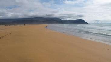 A vast, empty sandy beach stretches along the shoreline, with gentle waves meeting the coast. Distant mountains are visible under a partly cloudy sky, creating tranquility.