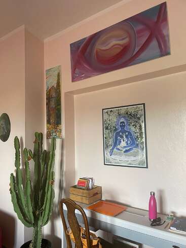A imagem retrata um canto de uma sala com uma mesa branca, um cacto e obras de arte nas paredes, apresentando uma atmosfera serena e criativa.