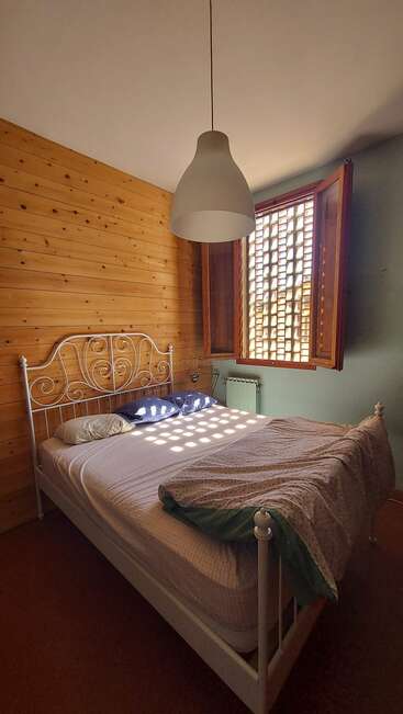 A imagem mostra um quarto aconchegante com uma estrutura de cama de metal branco, uma janela com persianas de madeira e uma luminária pendurada no teto, banhada pela luz natural do sol.