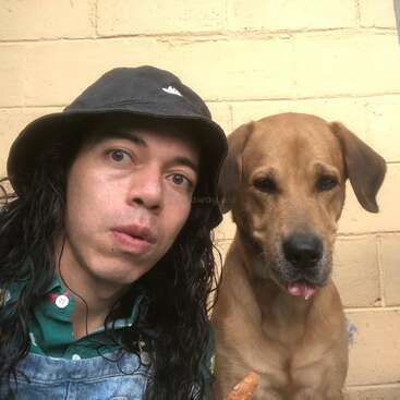 Une personne aux longs cheveux bouclés, portant un chapeau noir et une chemise verte, pose pour un selfie à côté d'un chien marron contre un mur de briques jaunes.