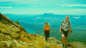 Two hikers walk up a rocky mountain trail, surrounded by green hills and a distant volcano, under a bright blue sky, enjoying the scenic view.