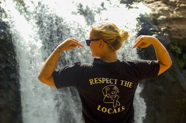 Eine Person mit blonden Haaren, die eine Sonnenbrille und ein schwarzes T-Shirt mit der Aufschrift "RESPECT THE LOCALS" trägt, steht vor einem wunderschönen Wasserfall und zeigt auf ihr T-Shirt.