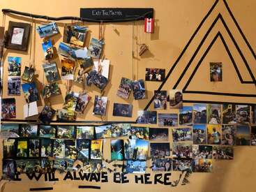 A collage-covered wall displays numerous colorful photographs of people, adventures, and memories. Black tape spells "I WILL ALWAYS BE HERE." Geometric lines and "Exit The Matrix" accentuate the décor.