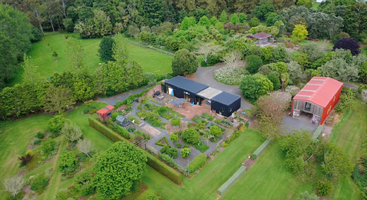 An aerial view of a lush, green property featuring two buildings, manicured gardens, pathways, expansive lawns, hedges, and surrounding dense trees creating a serene landscape.