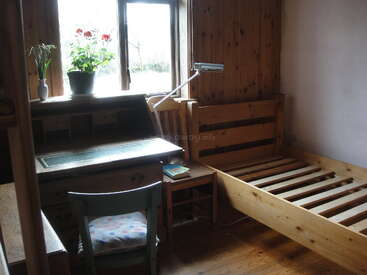 A small wooden bedroom features an empty single bed frame, a desk with a green chair, a lamp, a nightstand, and potted plants on the windowsill.