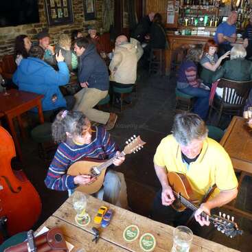 The image depicts two individuals playing guitars at a wooden table, surrounded by drinks and small toys, in a dimly lit room with other people seated around them.