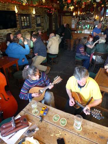 Das Bild zeigt zwei Personen, die an einem Holztisch Gitarre spielen, umgeben von Getränken und kleinen Spielzeugen, in einem schwach beleuchteten Raum, in dem noch andere Leute um sie herum sitzen.