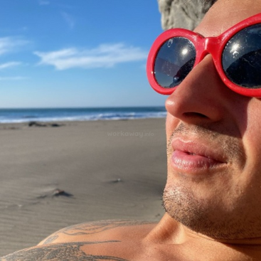 Una persona con grandes gafas de sol rojas se relaja en una playa de arena, disfrutando del tiempo soleado y del cielo azul despejado, con el océano de fondo.