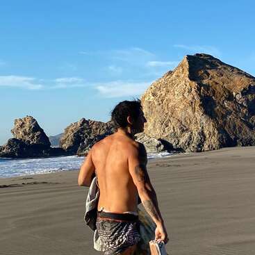 Un homme torse nu marche sur une plage de sable en tenant ses chaussures. De gros rochers et des vagues se trouvent à l'arrière-plan, sous un ciel bleu vif.