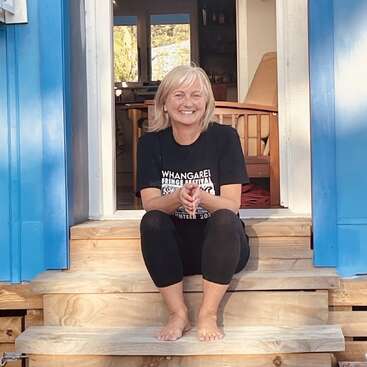 A smiling woman with blonde hair sits barefoot on wooden steps, wearing a “Whangarei Fringe Festival” t-shirt and black leggings, framed by bright blue doorways.