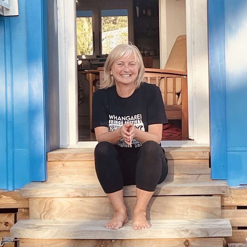 Une femme souriante aux cheveux blonds est assise pieds nus sur des marches en bois, vêtue d'un t-shirt « Whangarei Fringe Festival » et d'un legging noir, encadrée par des portes d'un bleu vif.