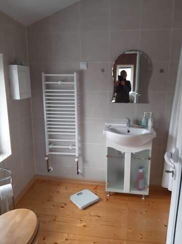 A tidy bathroom with light wood floor, white sink cabinet, wall mirror, towel warmer, scale, soap dispenser, and cleaner bottle under the sink. Person in mirror.