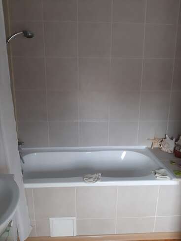 A clean, modern bathroom features a white bathtub with folded towels, seashell and starfish decorations, beige tiled walls, a showerhead, and a small sink partially visible.