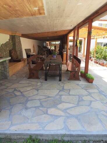 This image shows a covered patio with a stone floor, wooden benches, a long table, plants, rustic decor, and a cozy, inviting atmosphere for gatherings.