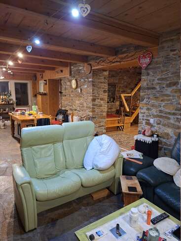This cozy rustic living room features stone walls, wooden beams, two sofas, a cluttered coffee table, and heartfelt decor with hanging hearts and homey touches throughout.