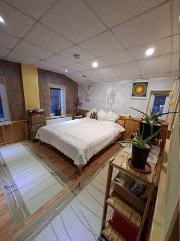 A cozy bedroom features a neatly made bed with a crocheted blanket, soft lighting, plants, dreamcatcher, artwork, wooden furniture, and warm carpets enhancing its inviting atmosphere.