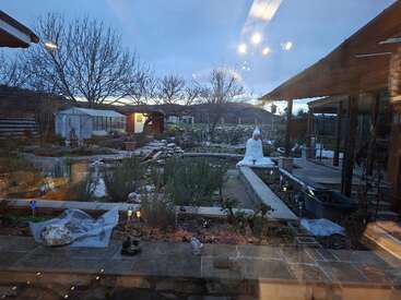 A peaceful garden scene at dusk with bare trees, a white Buddha statue beside a pond, greenhouses, lights reflecting off glass, and cozy wooden buildings surrounding.