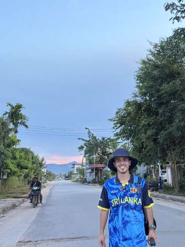Um jovem sorridente vestindo uma camisa azul do Sri Lanka e chapéu preto está em uma rua tranquila ao pôr do sol, com árvores, motocicletas e montanhas atrás.