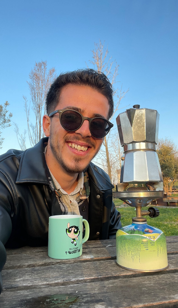A smiling man wearing sunglasses sits outdoors at a wooden table, enjoying coffee from a green cartoon mug, with a moka pot on a camping stove.