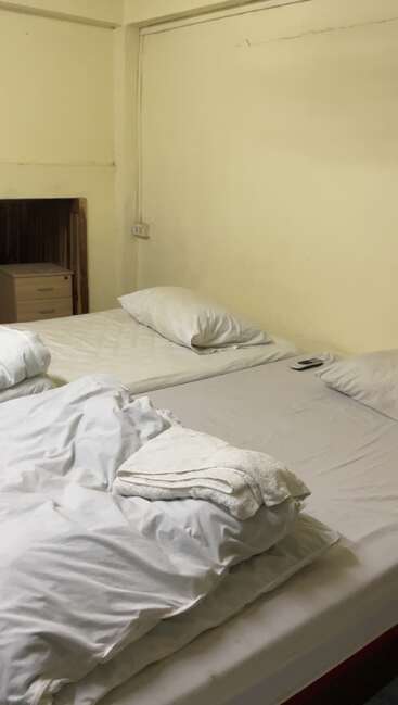 Esta imagem mostra um quarto simples com duas camas de solteiro, roupa de cama branca, dois travesseiros, toalhas dobradas, uma mesa de cabeceira e uma parede lisa de cor creme.