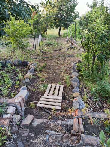Ein rustikaler, mit Steinen gesäumter Gartenweg führt unter einem Metallbogen hindurch. Überwucherte Pflanzen umgeben eine behelfsmäßige Brücke aus Holzpaletten und überall gibt es Bäume und Grünzeug.