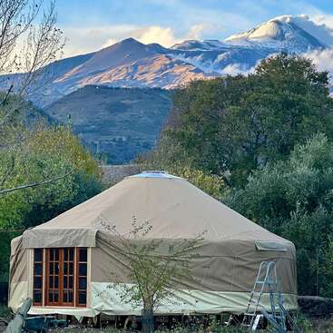 Zwischen grünen Bäumen steht eine Jurte mit einer Holztür und einer kleinen Leiter im Freien. Im Hintergrund erheben sich majestätische, teilweise schneebedeckte Berge.