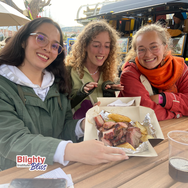 Drei Freunde sitzen an einem Tisch im Freien, lächeln und zeigen aufgeregt ein Tablett mit gegrilltem Fleisch und Kartoffelspalten. Getränke und legere Kleidung vervollständigen die Szene.