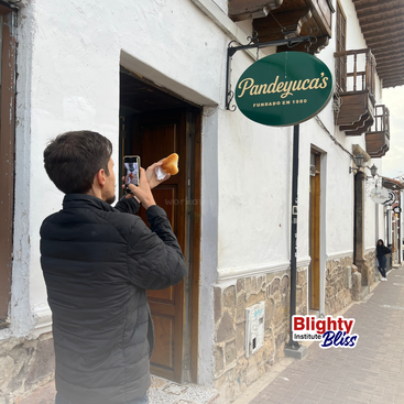 Ein Mann in einer schwarzen Jacke fotografiert ein Gebäck vor der Bäckerei "Pandeyuca's". Die Straße ist geprägt von weißen Wänden, Steindetails und charmanten Balkonen.