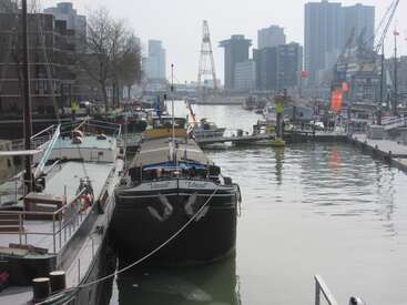 A boat named “Ideal” is docked in a busy harbor. Surrounding buildings, cranes, and other boats reflect an urban waterside cityscape under a hazy sky.