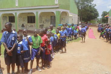 A group of school children lines up outside a green school building, wearing colorful uniforms. The atmosphere appears lively, cheerful, and organized on a sunny day.