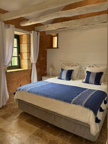 Cette chambre confortable présente des poutres apparentes rustiques, des murs en pierre et du carrelage. Un grand lit aux accents bleus et aux draps blancs est placé près de fenêtres à rideaux éclairées par le soleil.