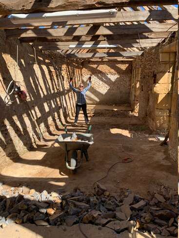 Une femme souriante se tient dans un bâtiment en pierre partiellement démoli et sans toit, levant les bras triomphalement. La lumière du soleil passe à travers les poutres. Une brouette, des pierres et des gravats remplissent le sol en terre battue.