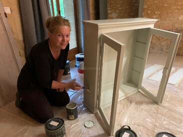 Une femme est agenouillée sur le sol et sourit en peignant une armoire blanche. Elle est entourée de pots de peinture et de pinceaux. La pièce a des murs en pierre et des portes en bois.