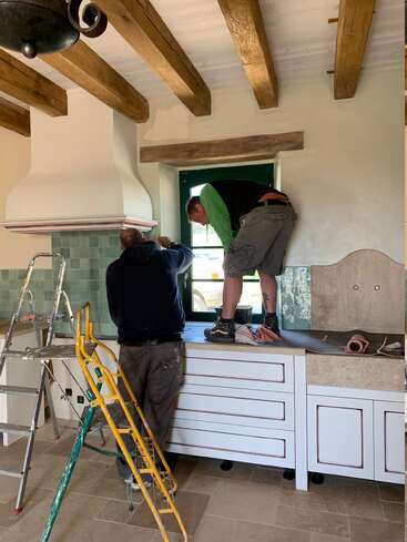 Deux hommes travaillent dans une cuisine rustique, l'un debout sur les comptoirs près d'une fenêtre, l'autre sur le sol. Des échelles et des outils se trouvent à proximité. Poutres du plafond visibles.
