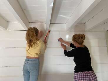 Deux femmes travaillent ensemble sur un mur en bois blanc, appliquant du calfeutrage et lissant le mur. Elles sont concentrées et collaborent au projet de rénovation de la maison.