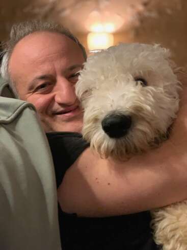 Un homme souriant serre affectueusement un chien blanc et duveteux dans ses bras. L'arrière-plan est doucement éclairé, créant une atmosphère chaleureuse et confortable, mettant en valeur leur lien étroit et heureux.