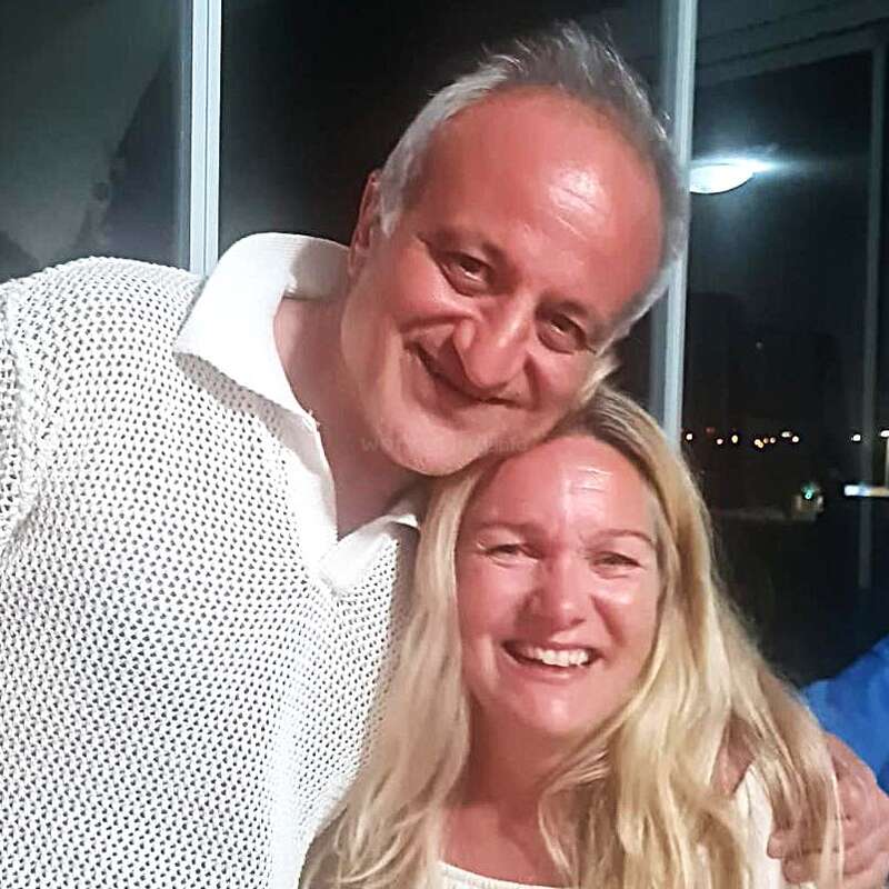 Un homme souriant aux cheveux gris enlace une femme blonde joyeuse. Ils semblent heureux et détendus, debout ensemble à l'intérieur, la nuit, avec les lumières de la ville en arrière-plan.