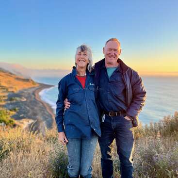 Um casal sorridente está junto numa colina gramada com vista para um lindo litoral ao pôr do sol, curtindo a vista panorâmica do mar e do céu dourado, bem agasalhados.