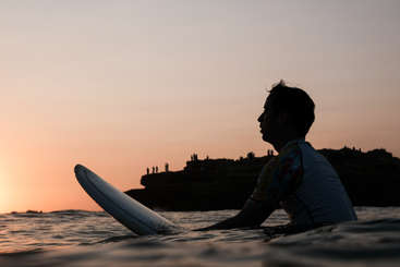 Das Bild zeigt eine Person, die am Ufer steht und auf den Ozean hinausblickt, mit einem Surfbrett im Hintergrund, vor dem Hintergrund einer ruhigen Strandszene.