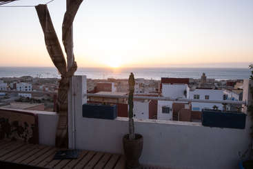 Das Bild zeigt eine ruhige Außenszene eines Gebäudes mit einem Balkon mit Geländer, einem Fenster und einer Tür vor dem Hintergrund eines klaren Himmels und eines Gewässers.