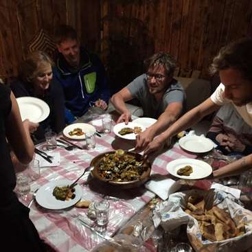 The image depicts a group of people gathered around a table, enjoying a meal together, with a large bowl of food and plates of food on the table.