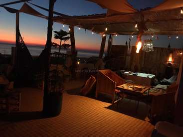 Das Bild zeigt eine ruhige Dachterrasse bei Sonnenuntergang mit Tisch, Stühlen und Lichterketten und einem atemberaubenden Meerblick im Hintergrund.