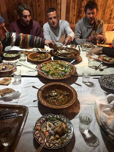 Das Bild zeigt eine Gruppe von Männern, die vor einer warmen Holzwand an einem Tisch sitzen, der mit einer Auswahl an Speisen beladen ist.